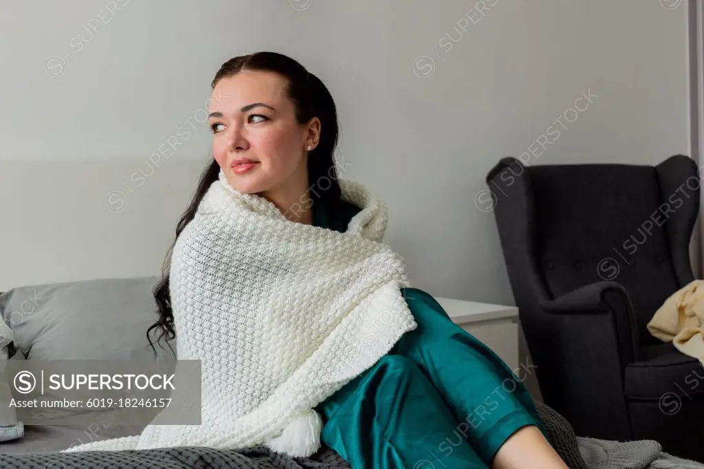 Young woman is wrapped in white plaid while sitting on bed in bedroom.