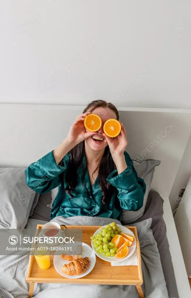 Young woman laughs and holds oranges near her eyes. Home comfort zone and wellness concept.