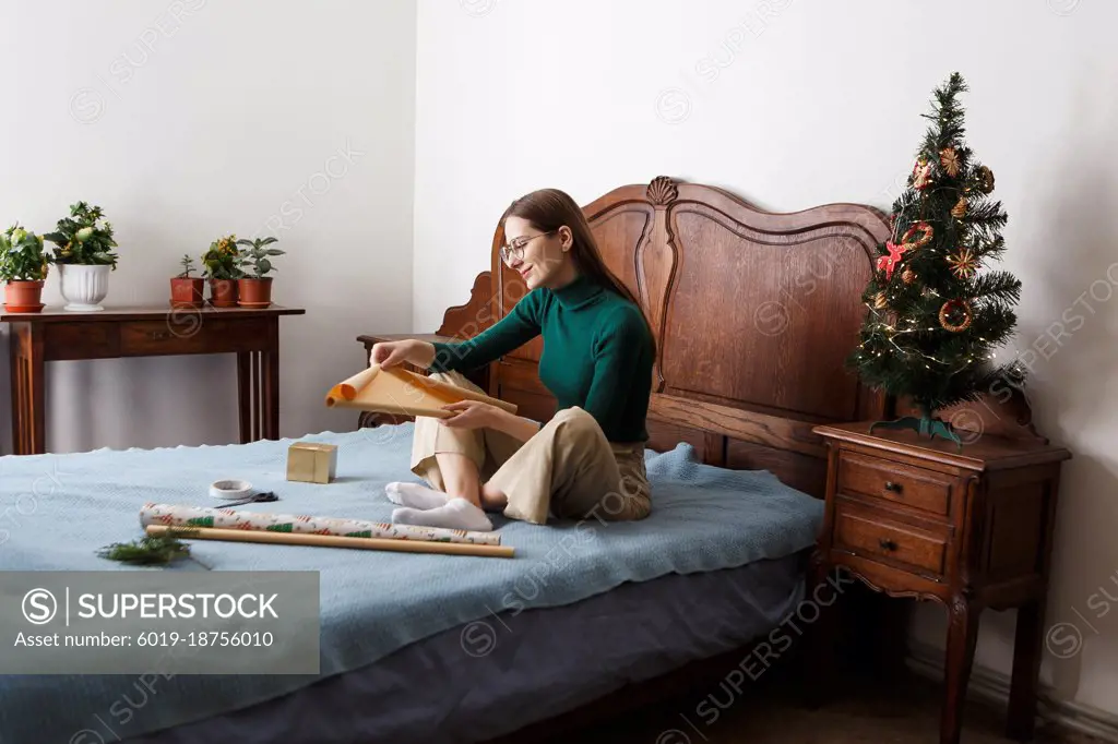 young woman packs a Christmas present while sitting on the bed at home