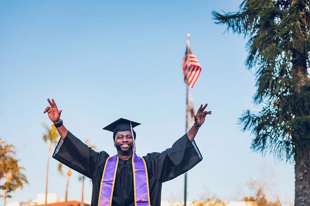 Young man wearing a graduation gown, american flag in the background.