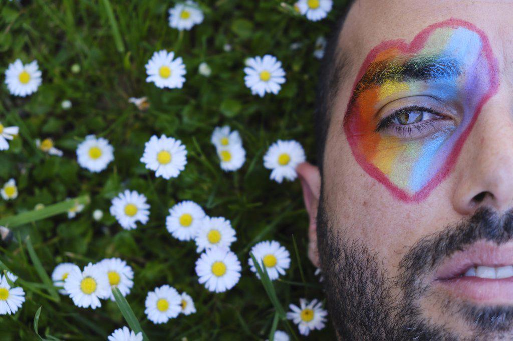 gay man with green eyes and beard lying on a lawn full of daisies, with a heart painted on his eye in the colors of the lgtb flag. pride