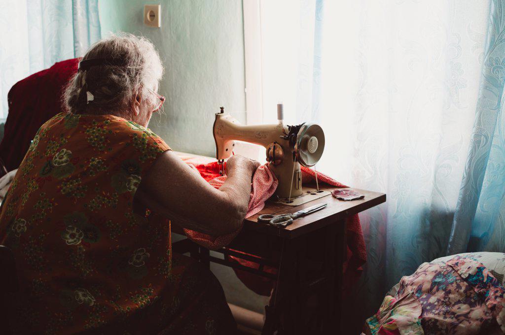 Old gray-haired woman with curly hair sews on retro sewing machine