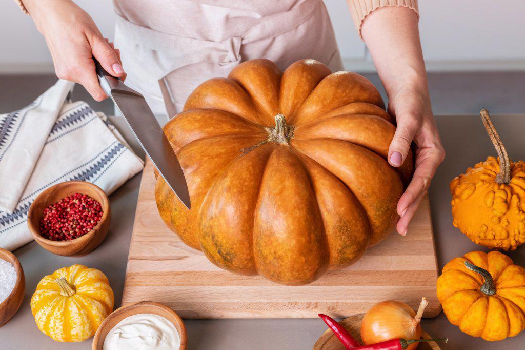 A woman in the kitchen cuts a large pumpkin with a knife.