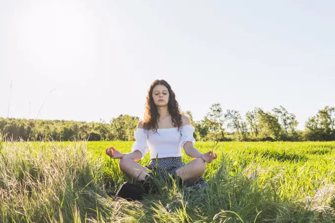 woman meditating in the middle of a green wheat field against the sun