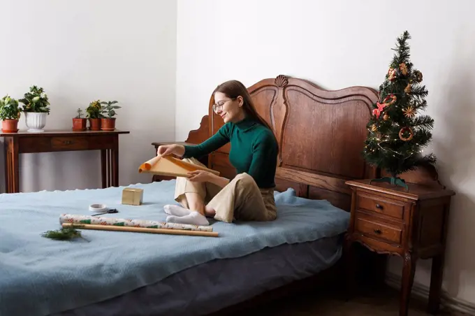 young woman packs a Christmas present while sitting on the bed at home