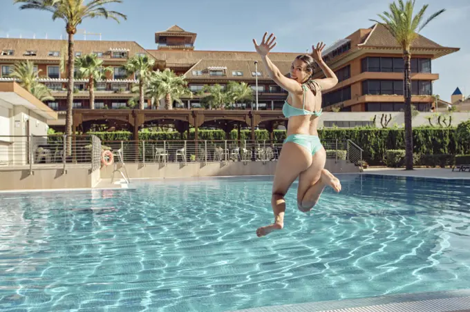 Excited female jumping in pool at resort against sunny sky