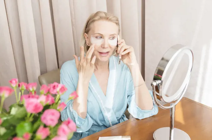 A mature woman with eye mask is talking on the phone at home.