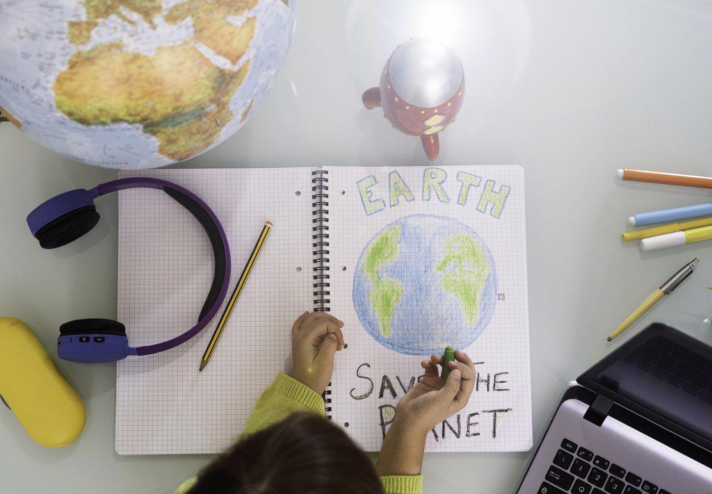 Smiling child girl draws planet earth with wax colors on school notebook for Earth day - Little activist girl writes the message Save the Planet - Protection of environment, global warming - Top view